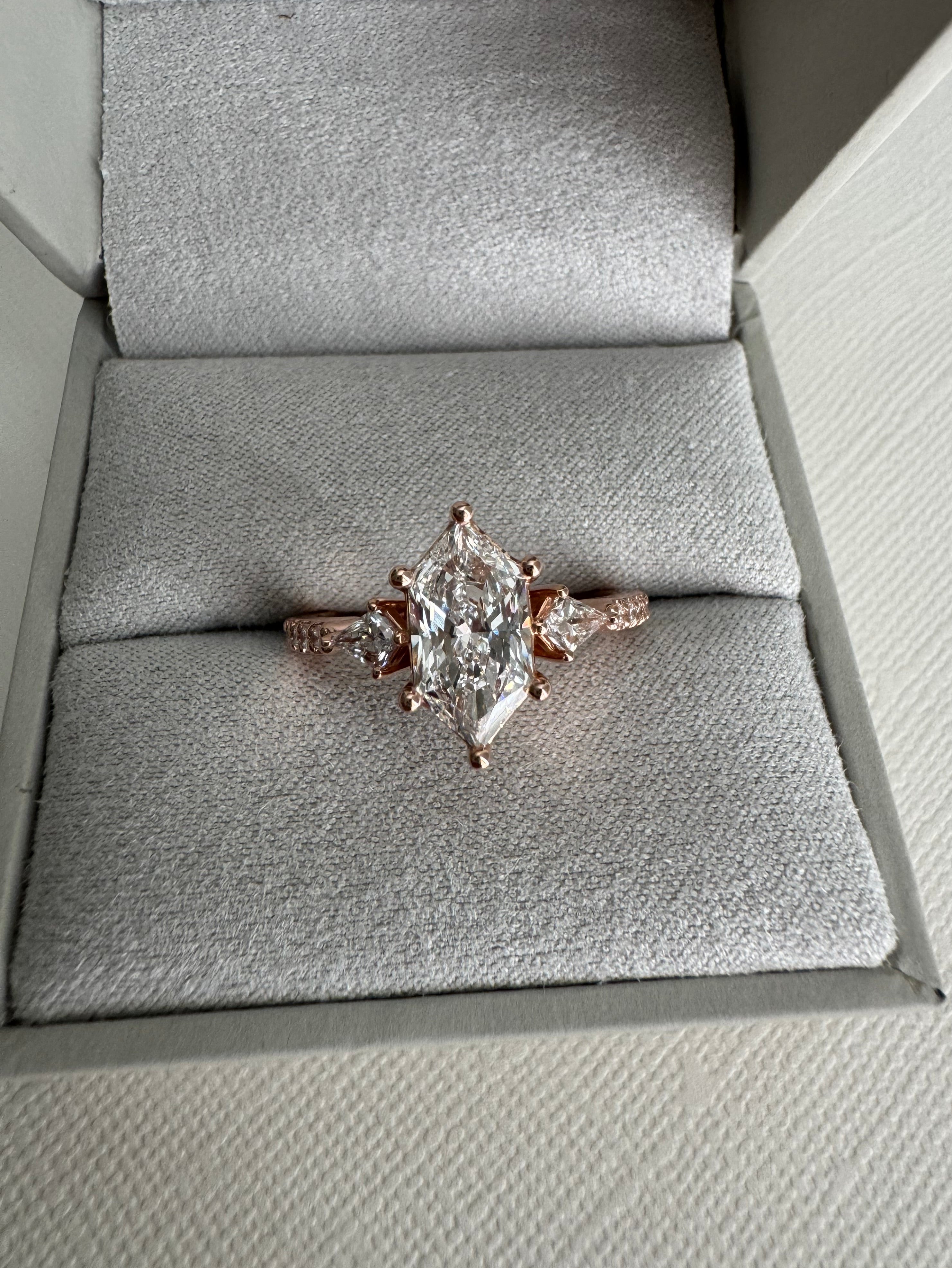 Custom 14k rose gold lab grown diamond engagement ring. Center= 1.69 ct lab grown elongated hexagon. Accents are 2 lab grown kite diamonds (0.29 cttw), 12 lab grown round diamonds (approx 0.18 cttw), 1 round emerald, 1 round tsavorite garnet. Size 8.5.