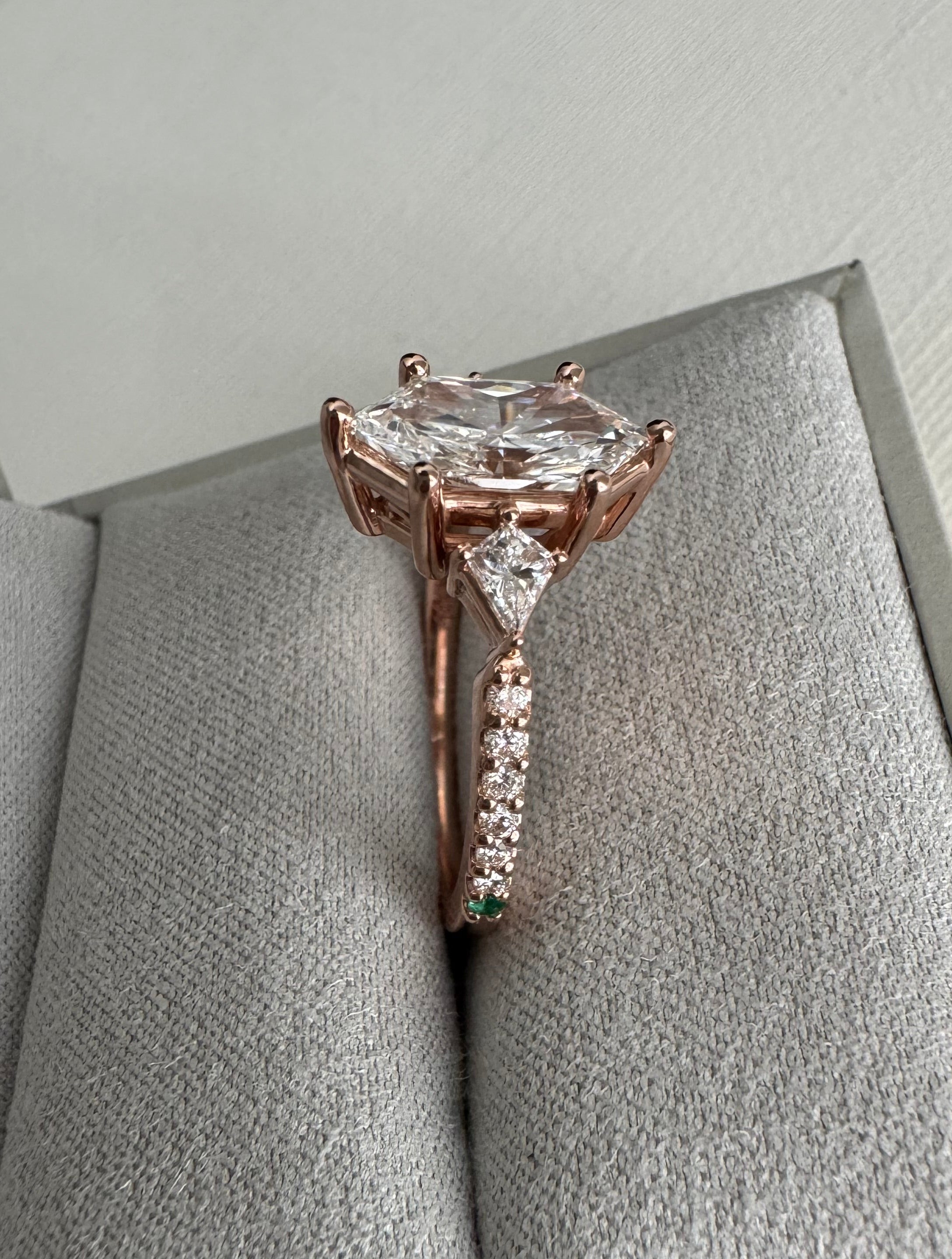 Custom 14k rose gold lab grown diamond engagement ring. Center= 1.69 ct lab grown elongated hexagon. Accents are 2 lab grown kite diamonds (0.29 cttw), 12 lab grown round diamonds (approx 0.18 cttw), 1 round emerald, 1 round tsavorite garnet. Size 8.5.